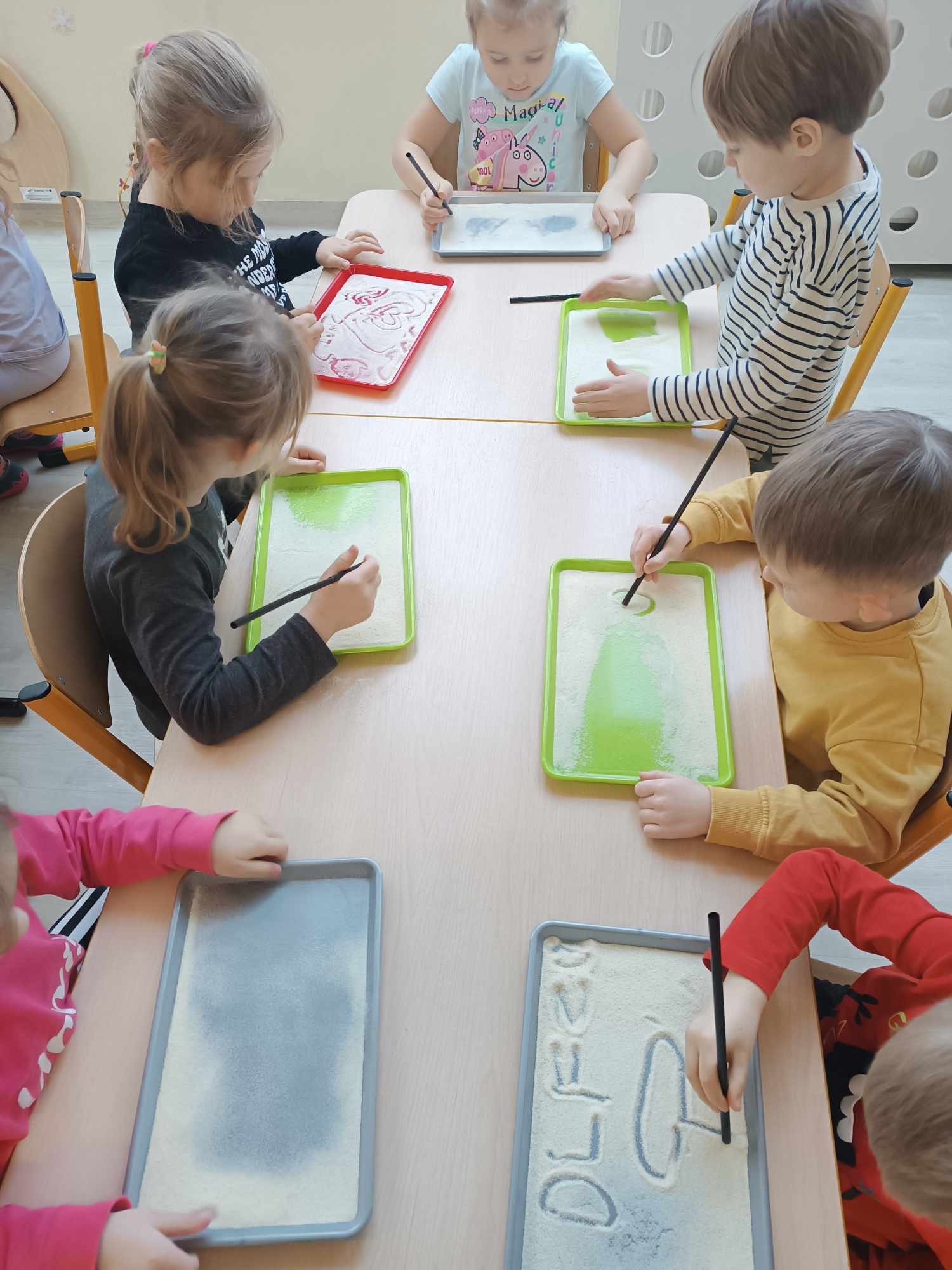 dzieci przy stolikach rysują na kaszy słomką